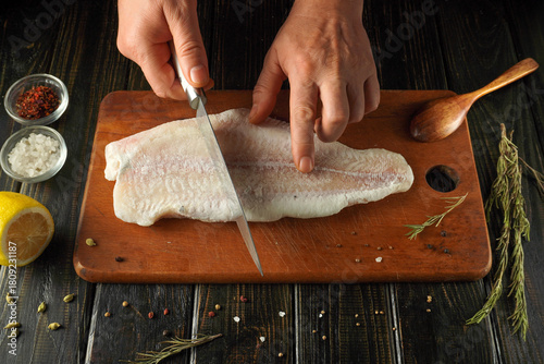Hands skillfully slice a fresh fish fillet on a wooden cutting board. Nearby, bowls hold sea salt, spices, and a lemon, ready for seasoning and cooking