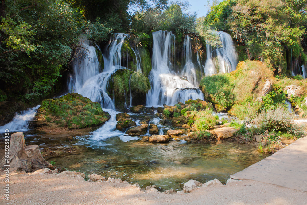 Fototapeta premium Lush Kravice Waterfalls cascade into emerald water, surrounded by dense greenery near Medjugorje, Bosnia—an idyllic natural landscape with vibrant mist and serene reflections.