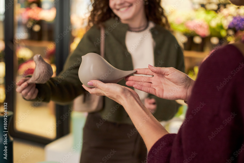 Obraz premium Caucasian young adult woman holding ceramic bird figurine while smiling at another Caucasian young adult woman in flower shop, colorful floral arrangements visible in background