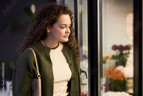Young adult Caucasian woman standing indoors looking thoughtfully at flower arrangements behind glass display case, curly hair framing face, holding handbag over shoulder Free Download Young adult Caucasian woman standing indoors looking thoughtfully at flower arrangements behind glass display case, curly hair framing face, holding handbag over shoulder - GFXLoad