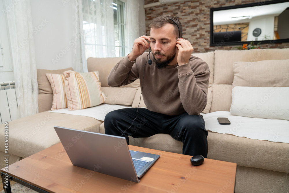 Fototapeta premium Man setting up headset, preparing for remote video conference call from home office couch, modern telecommuting lifestyle