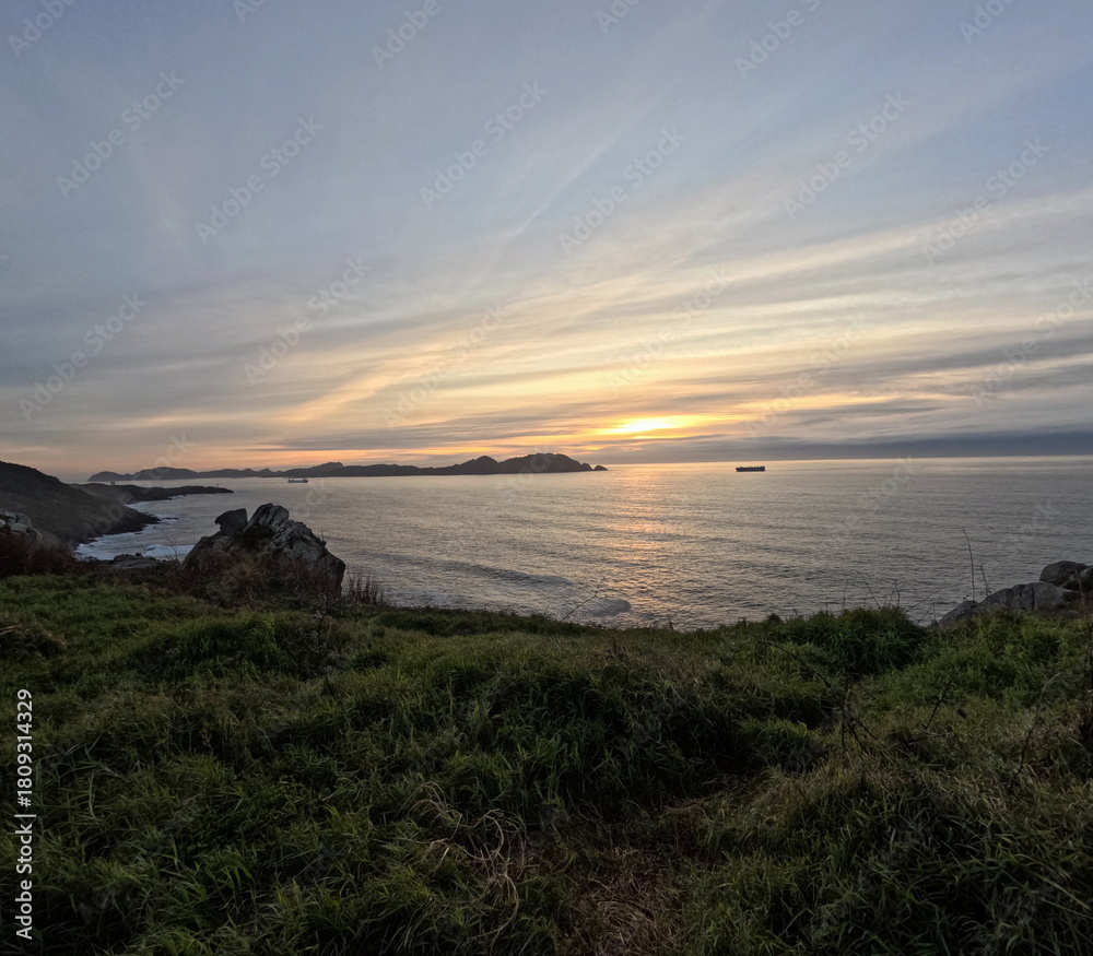 Obraz premium Golden sunset over the Atlantic Ocean from Cabo Home, Galicia, Spain The Cíes Islands silhouette in the background and a cargo ship sailing under a sky filled with soft clouds 