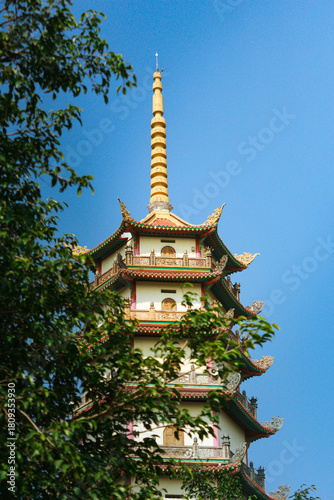 A tall pagoda tower with intricate traditional architecture rising above green foliage under a clear blue sky. A peaceful scene highlighting Asian cultural and religious design.
