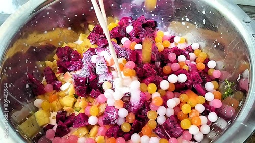 Preparation of a tropical fruit dessert: Coconut milk poured over mixed tropical fruit and colorful jelly pearls in a stainless steel container