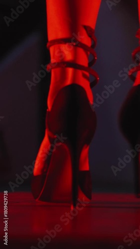 Vertical close-up of a woman legs turning in black high-heeled shoes with straps and a black dress.indoors at night with a red light surrounding and illuminating 