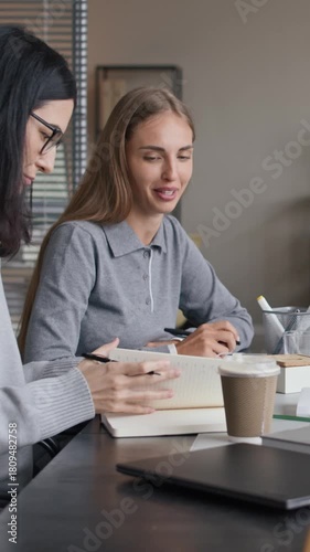 Vertical medium shot of young Caucasian female colleagues discussing business project sitting at conference table during meeting in modern office
