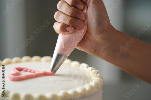 Person's hand decorating a white cake with pink buttercream heart frosting, a sweet and delicate baking process in action