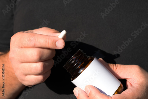 Hands present a capsule above an amber bottle with white label against dark fabric; bright sunlight, clean lines and negative space convey vitamin health and confident lifestyle.