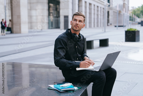 Thinking man with laptop and notebook in a sleek urban plaza, immersed in modern city life and productivity.