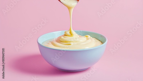 Creamy yellow custard pouring from wooden spoon into light blue ceramic bowl on pink background soft studio lighting
