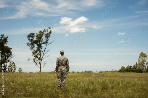 Male soldier standing with back to horizon in grassy field wearing camouflage uniform and cap