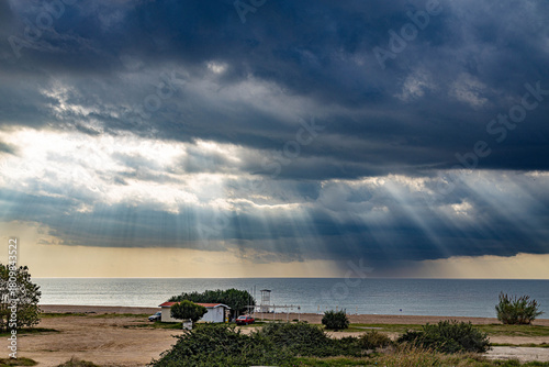 Fototapeta Naklejka Na Ścianę i Meble -  Serene autumn morning on a deserted Mediterranean beach Sunbeams break through gray clouds above a white house with tile roof, creating a dramatic light pattern over the sea. Sorgun, Antalya, Turkey