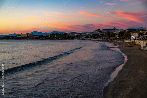 Fototapeta Naklejka Na Ścianę i Meble -  Serene evening view of the Mediterranean coastline in Side after sunset. A curved deserted beach with promenade, hotels among palms, and mountain silhouettes under a pink twilight sky. Antalya, Turkey