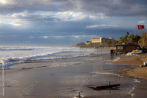 Fototapeta Naklejka Na Ścianę i Meble -  Powerful storm with massive waves crashing on a wet sandy beach under sunny skies. Wind bends palm trees and creates spray mist over the turbulent sea. Mediterranean coast, Side, Turkey.

