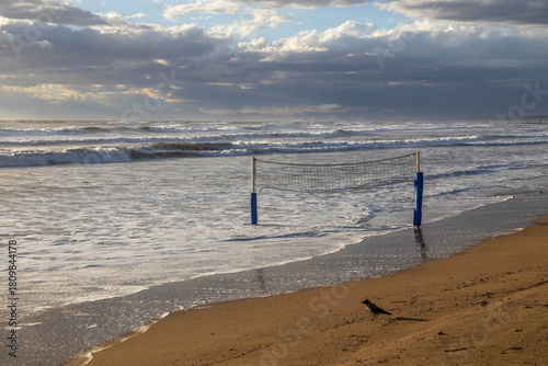 Fototapeta Naklejka Na Ścianę i Meble -  Powerful storm waves cover a beach under sunny skies, with volleyball poles standing in the surf and a crow on the sand. A dramatic scene of nature's force at a Mediterranean resort in low season.

