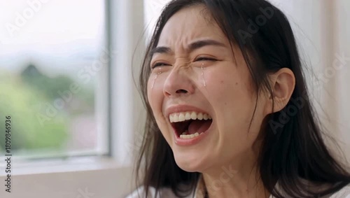 Young Asian woman laughing hysterically with mouth wide open, expressing joy and amusement in a bright indoor setting.