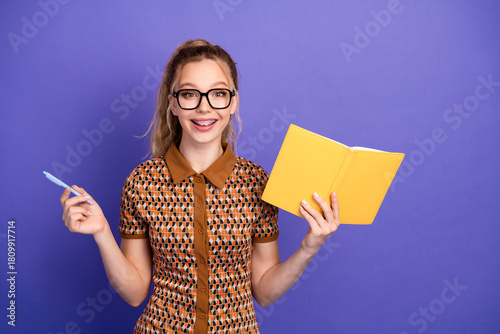Young girl student with glasses smiles holding a yellow notebook and pen against a purple background for education fashion lifestyle stock photo