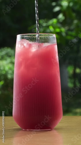 Red berry drink with ice cubes in a tall ribbed glass. Stirring refreshing soft drink in a glass outdoors. Vertical slow motion