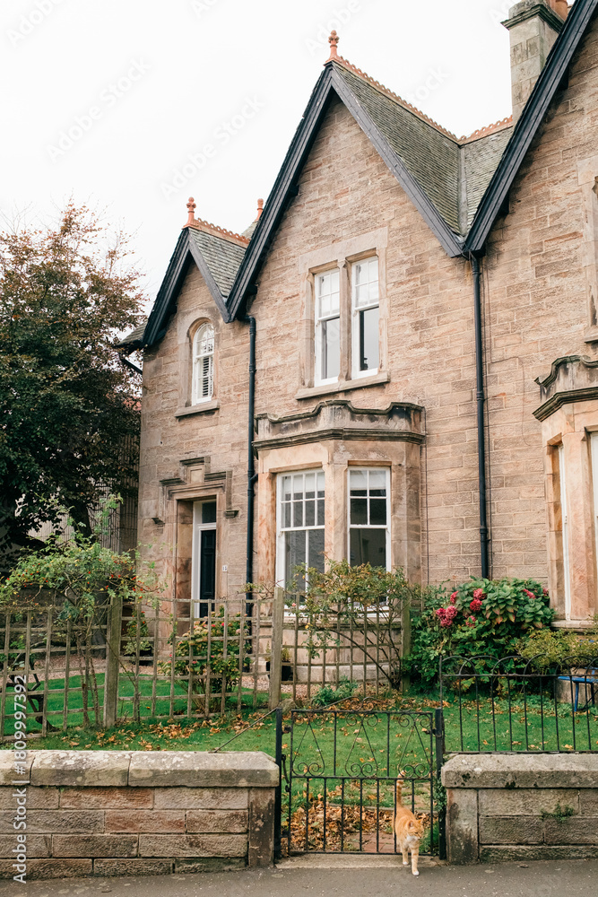 Obraz premium Traditional stone home with gabled roof, bay window, and front garden behind wrought iron gate.