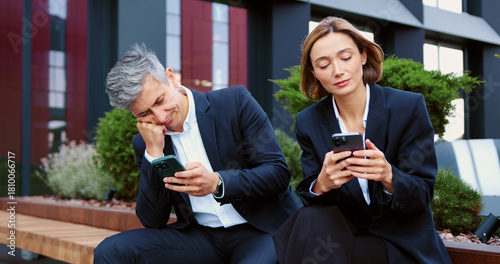Sad disinterested business man and woman looking disappointedly at uninteresting information on their smartphones while sitting outside on a modern office background. Disappointed business people in