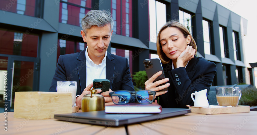 © serg - Sad disinterested business man and woman looking disappointedly at uninteresting information on their smartphones and drinking coffee during lunch break sitting on the summer terrace of the cafe. © serg - Sad disinterested business man and woman looking disappointedly at uninteresting information on their smartphones and drinking coffee during lunch break sitting on the summer terrace of the cafe.