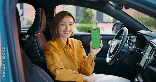 Fotografía Confident young businesswoman in casual attire sitting in a car, smiling and holding a smartphone with a green screen