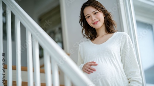 自宅の白い階段で手すりに手を添え微笑む白い服の妊婦