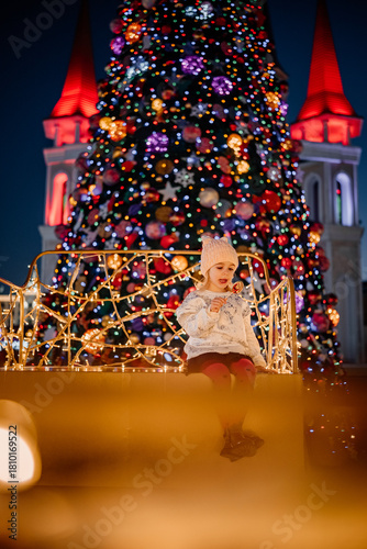 Child enjoying a lollipop in front of a beautifully decorated Christmas tree at night