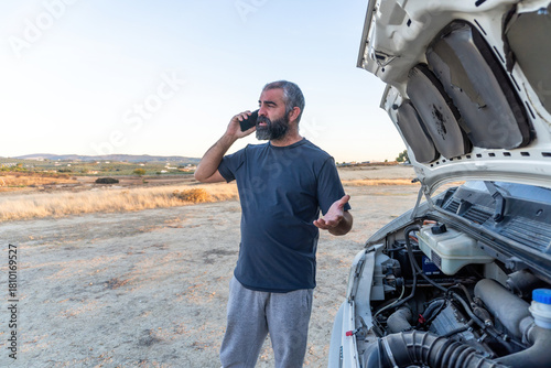 A man stands next to a camper van with the hood open, talking on his phone and gesturing as he seeks assistance in a remote rural area at sunset.