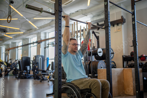 Disabled man in wheelchair doing vertical pull ups for spinal injury recovery using equipment in sports center on daily gym session. Male patient with legs immobility working on upper body strength.