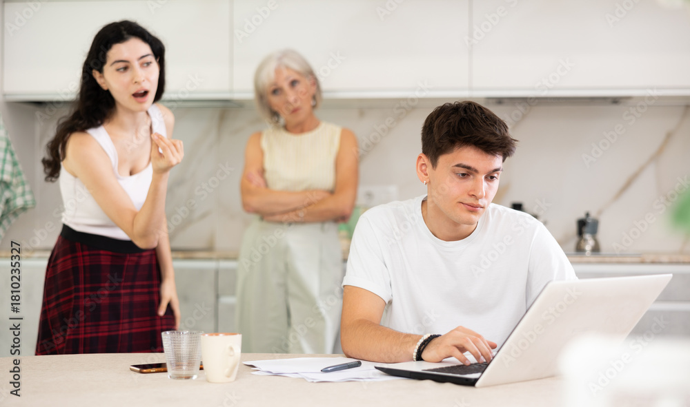 Obraz premium Young guy while working on laptop listens to claims of elderly mother and young sister girl in incompetence, unsuccessful study results