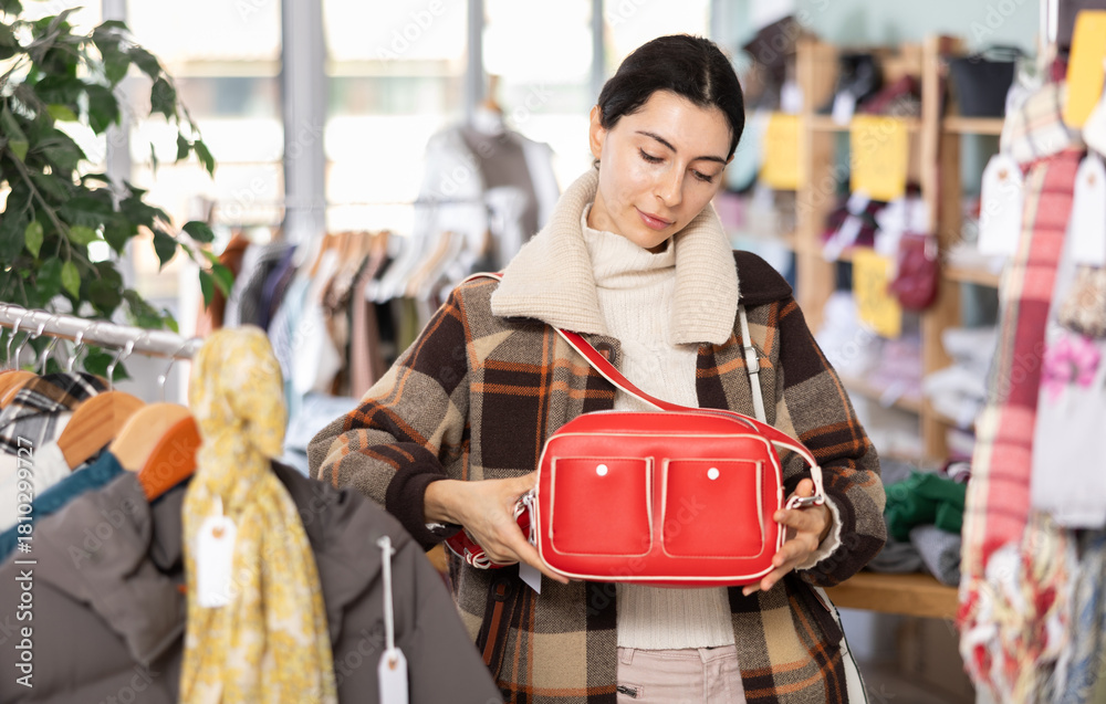 Obraz premium Sale of winter clothing collection. Positive young girl standing against background of shelves with clothes in store and choosing new bag