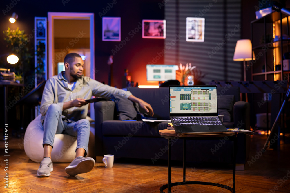 © DC Studio - African American freelancer studies candlestick charts on a laptop in his living room. Day trader monitoring stock exchange data, growth trends and financial figures while working remotely.