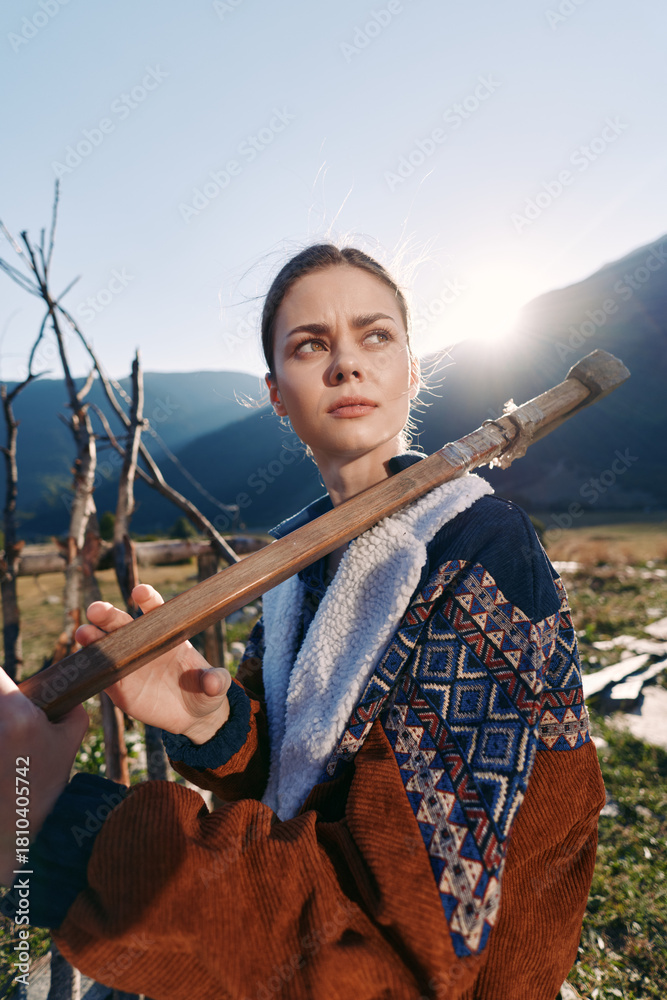 Naklejka premium Woman outdoors in mountains holding a wooden staff across her shoulders, rural portrait at sunrise with patterned jacket and meadow valley background, thoughtful gaze in warm sunlight.