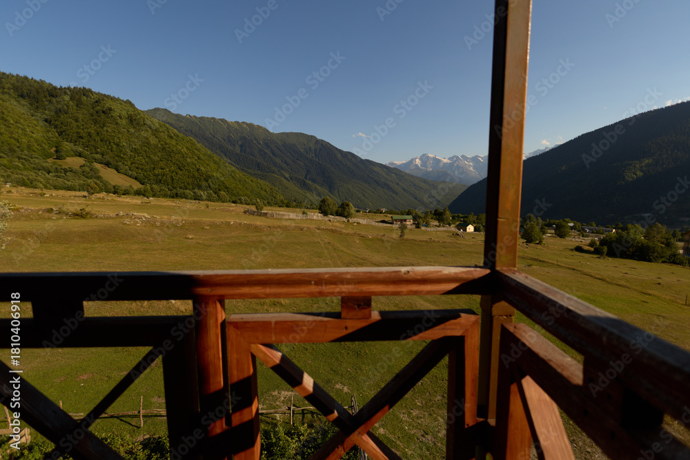 Obraz premium Balcony wooden railing overlooking mountains and valley with green meadow and countryside field, rustic cabin view under clear sky and distant snowy peaks for nature retreat.