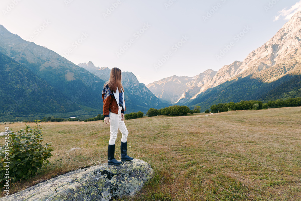 Naklejka premium Woman standing on rock in mountain valley hiking with boots and jacket, looking toward distant mountains. Landscape and nature scene for outdoor adventure, solo travel and exploration.