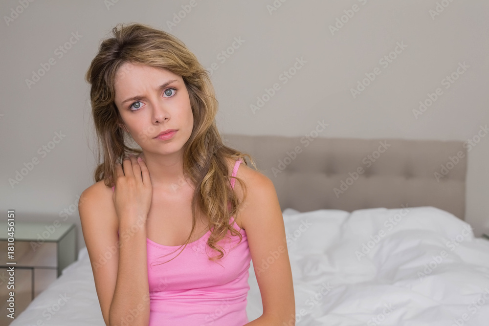 Naklejka premium Adult female sitting on bed, touching neck and looking toward camera in pink tank top
