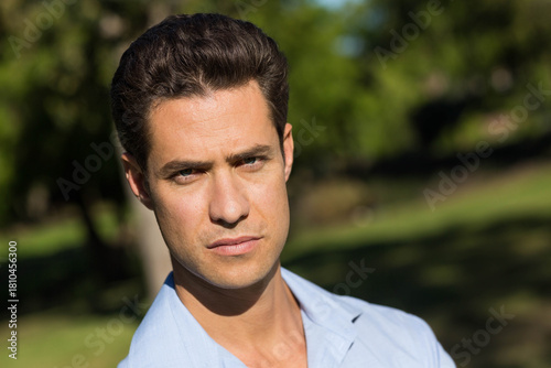 Mid-30s male standing posed in sunny park wearing light blue collared shirt with blurred background