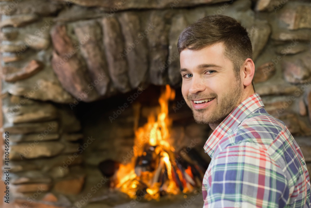 Naklejka premium Adult bearded man smiling, sitting by stone fireplace burning logs, wearing plaid shirt, copy space