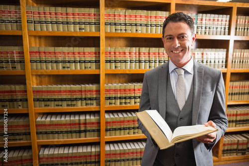 Man in suit standing by wooden bookshelves in law library holding open legal volume, copy space