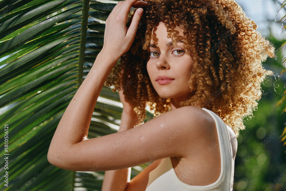 Naklejka premium Curly-haired woman posing outdoors in white top near tropical palm leaves with natural sunlight and green blurred background under warm summer conditions.