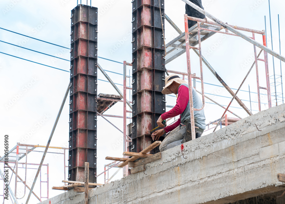 Naklejka premium A construction worker, wearing a long-sleeved shirt and a wide-brimmed sun hat, crouches on a newly poured concrete beam or slab.