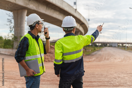 Two Construction Workers in Bright Safety Vests Communicating at Road Construction Site Under Bridge with Laptop and Walkie-Talkie Observing Work Progress
