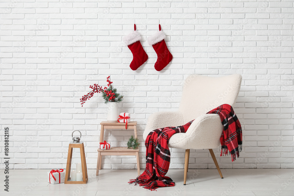 Obraz premium Interior of room with armchair, Christmas lantern, candles, gift boxes and red berries branches in vase on stepladder near white brick wall