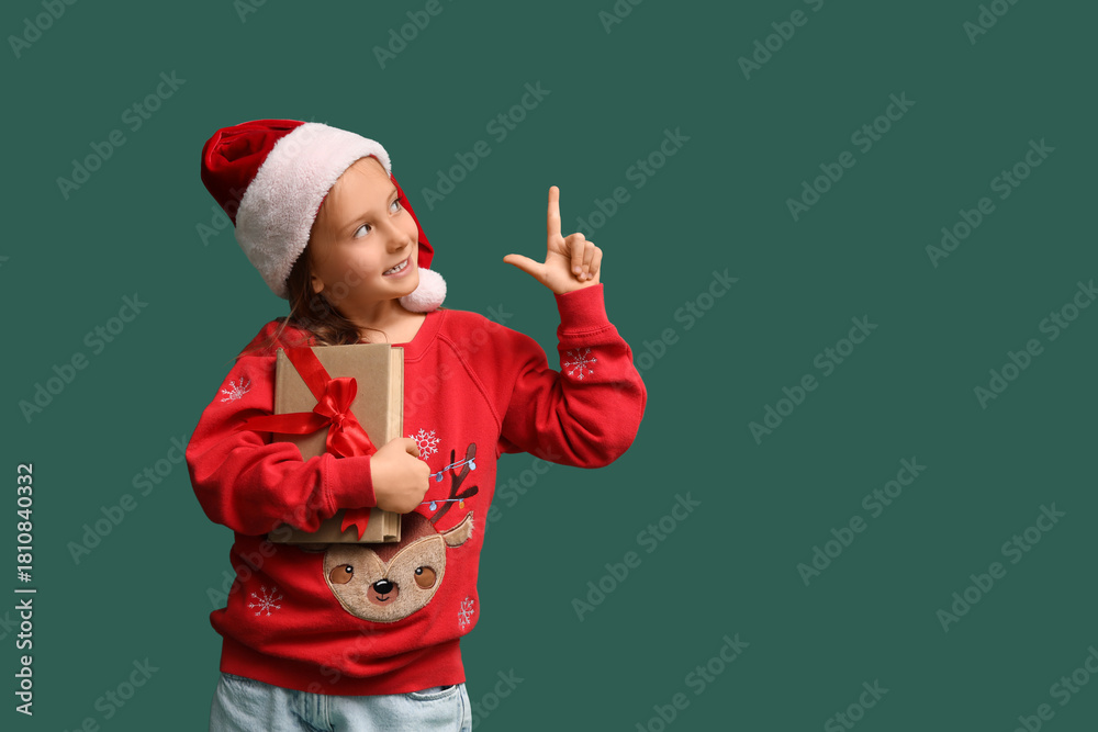 Obraz premium Cute little girl in Santa hat with gift book pointing at something on green background