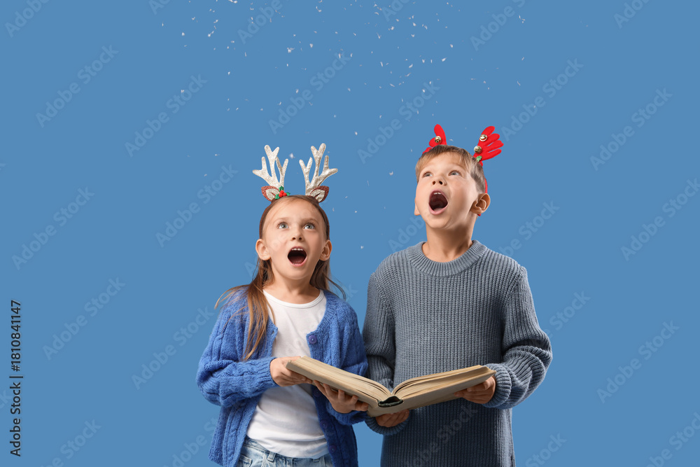 Fototapeta premium Surprised little children in Christmas reindeer horns with book and snow on blue background