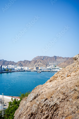 A bright coastal landscape in Muscat, Oman, featuring clear blue skies, turquoise water, rugged rocky mountains, and a line of modern white buildings along the waterfront
