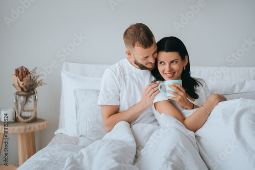 A cozy morning in bed with a couple sharing coffee under a white duvet, radiating warmth and affection in a minimalist setting.