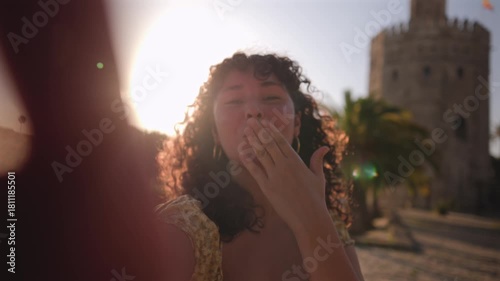 Happy young woman vlogging for social media, pointing at the torre del oro in seville, spain, smiling and blowing a kiss to the camera during a beautiful sunny day with a warm lens flare