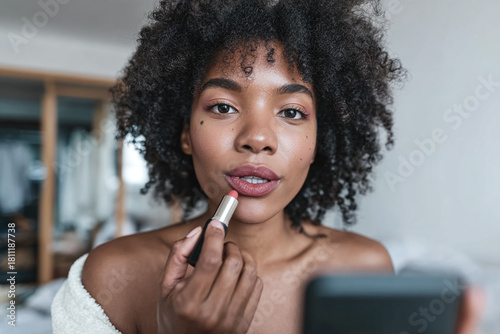Over-the-shoulder UGC shot of woman with medium brown skin and curly hair applying lipstick, phone on tripod visible, casual outfit, soft window light, shallow depth, realistic textures.